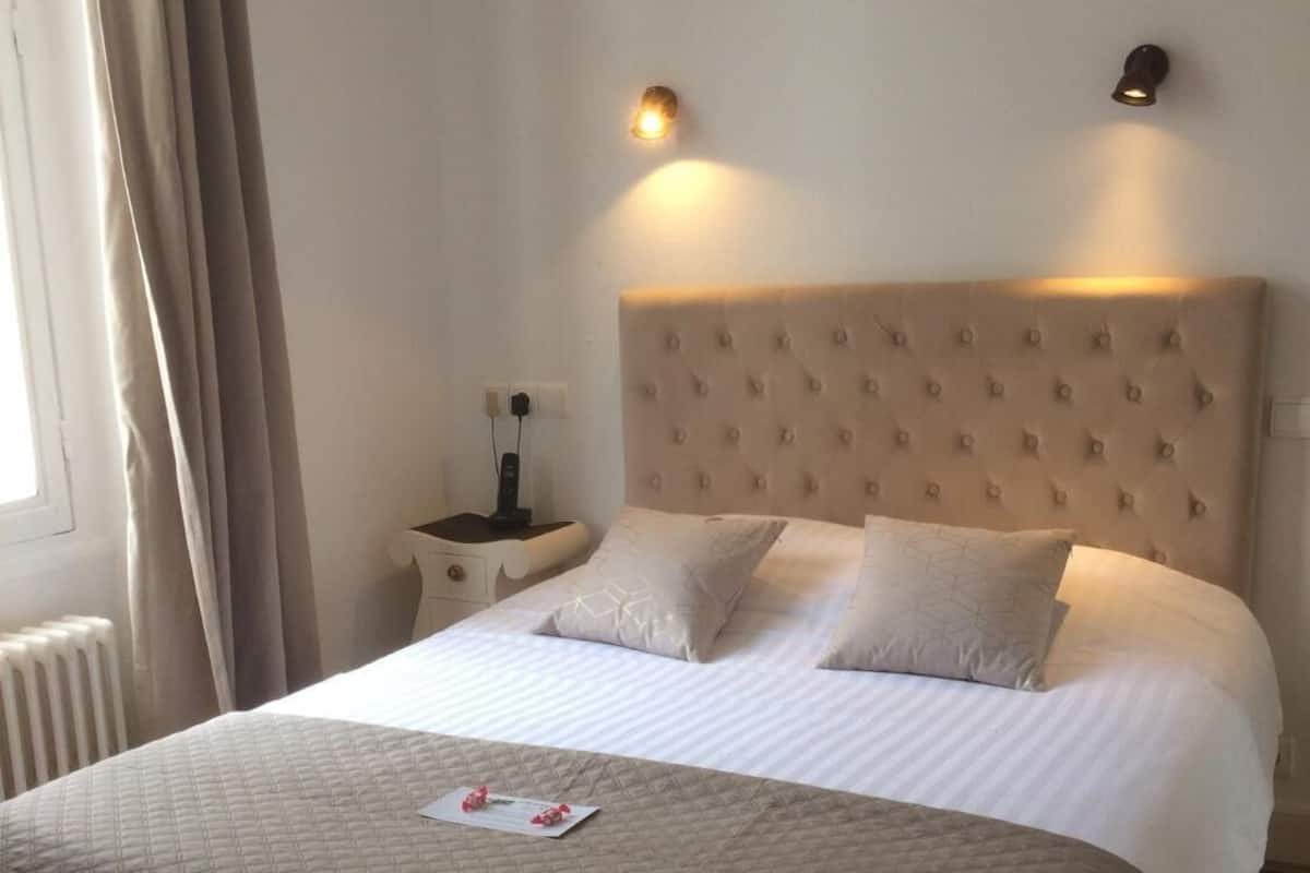 A neatly made bed with a quilted bedspread and two pillows, a bedside table with a lamp, and a wall-mounted light fixture.