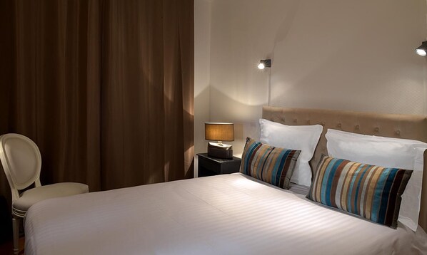 Habitación de hotel con una cama grande, una mesita de noche con lámpara, una silla y un ventanal con cortinas.