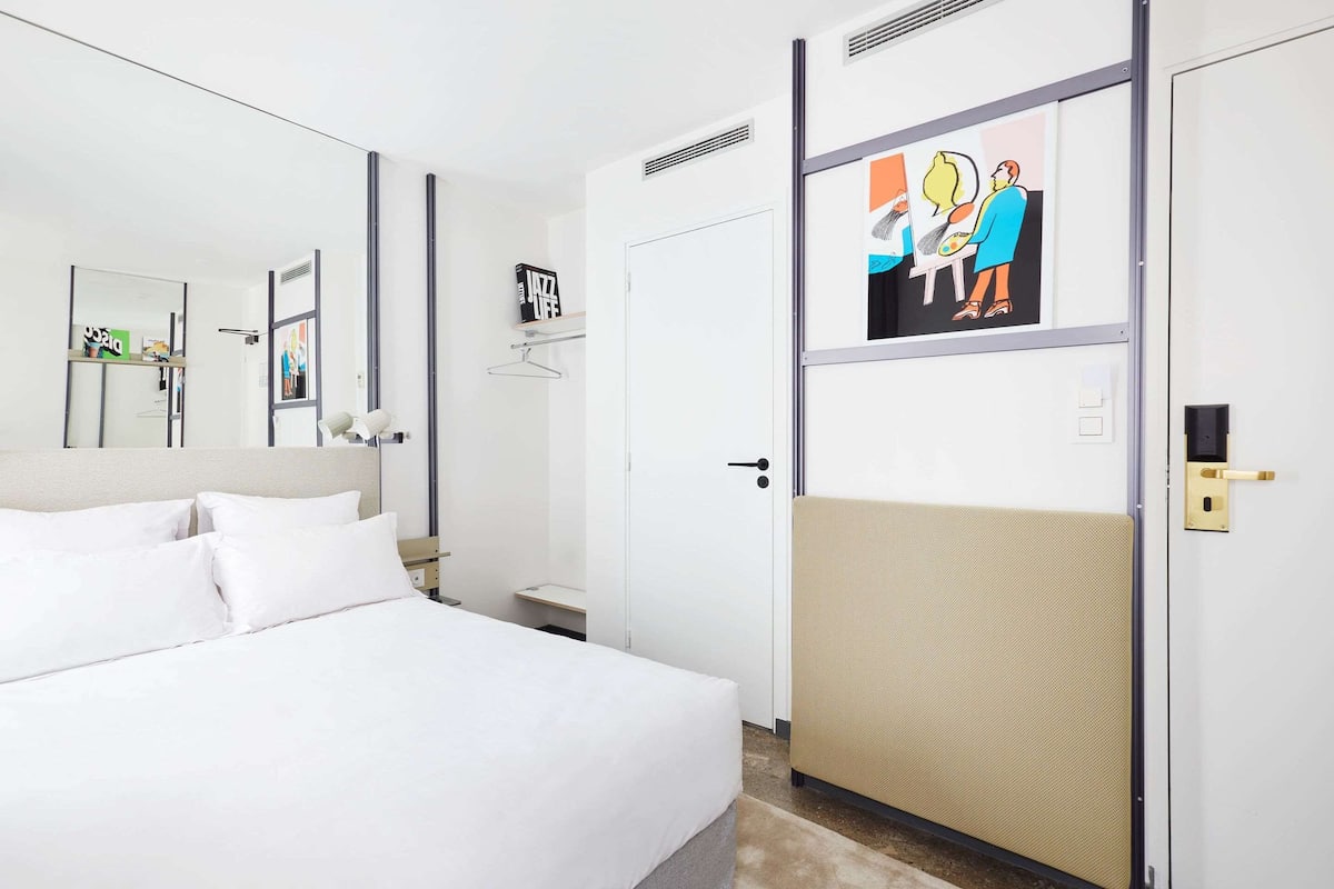 A hotel room with a bed, a wardrobe, a mirror, and a door with a colorful artwork.