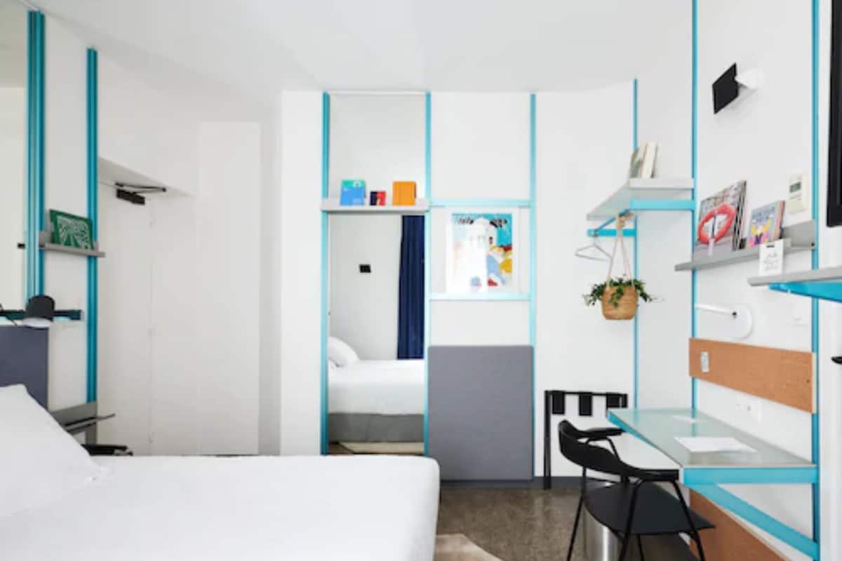 A modern hotel room with a bed, desk, chair, and a small kitchenette area.