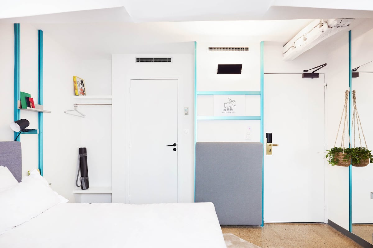 A modern hotel room with a bed, a desk, a hanging plant, and a door with a small window.