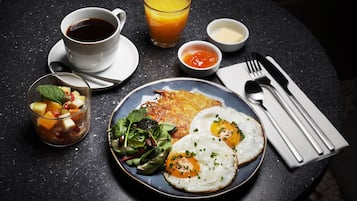Ein Frühstückstisch mit Eiern, Rösti und Obstsalat, einer Tasse Kaffee und einem Glas Orangensaft.
