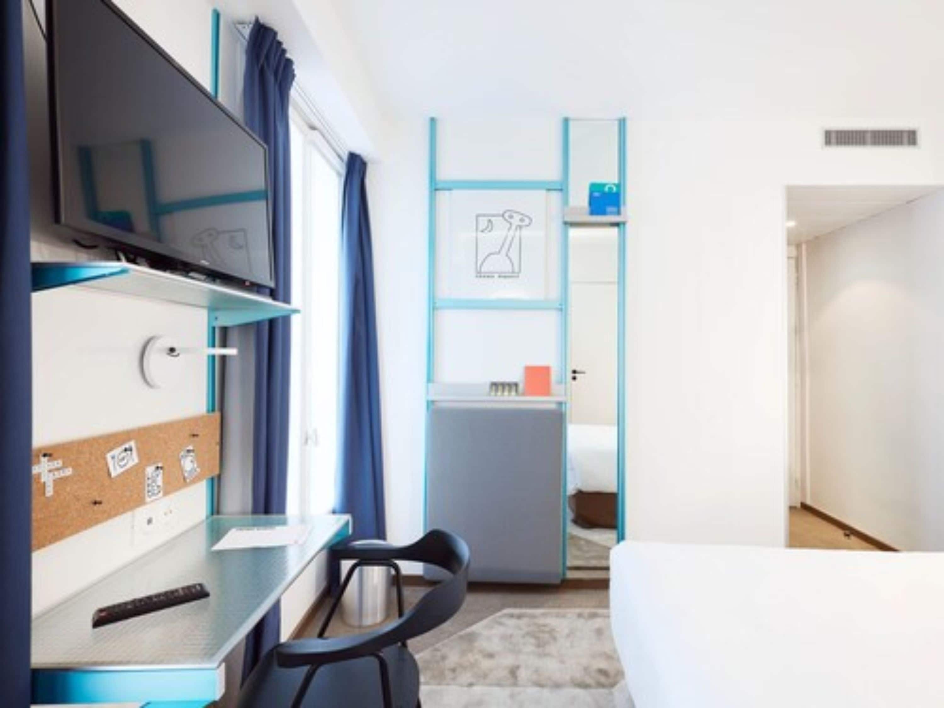 A compact hotel room with a bed, a desk with a chair, a TV, and a small kitchenette.