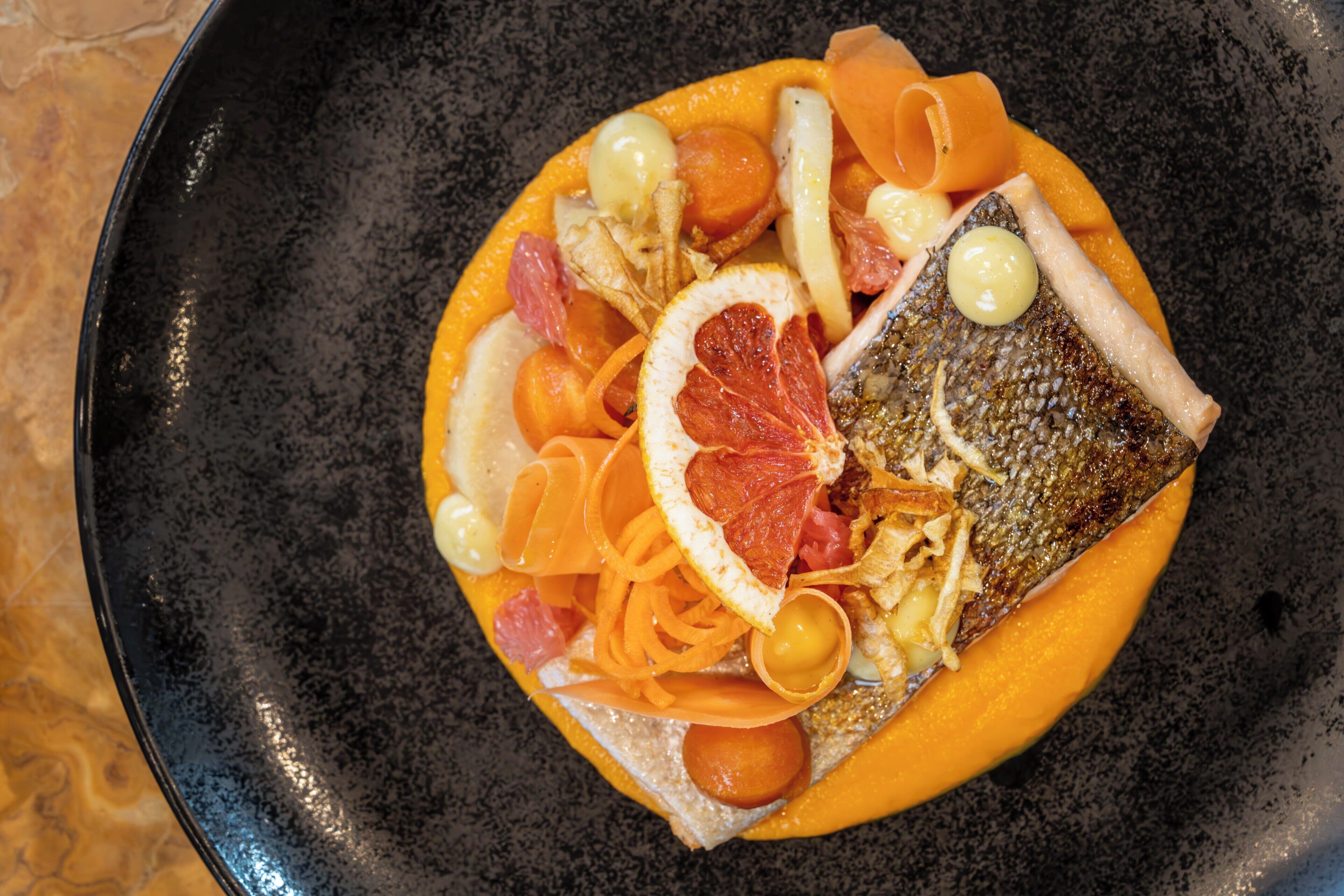 A plate with a fish, vegetables, and a slice of orange on a black plate.