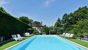 Una piscina rectangular con sillones de descanso a ambos lados, rodeada de vegetación y una casa al fondo.