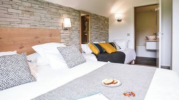 A hotel room with a stone wall, a wooden headboard, a bed with patterned pillows, a bedside table with a plate of food, and a bathroom visible through an open door.