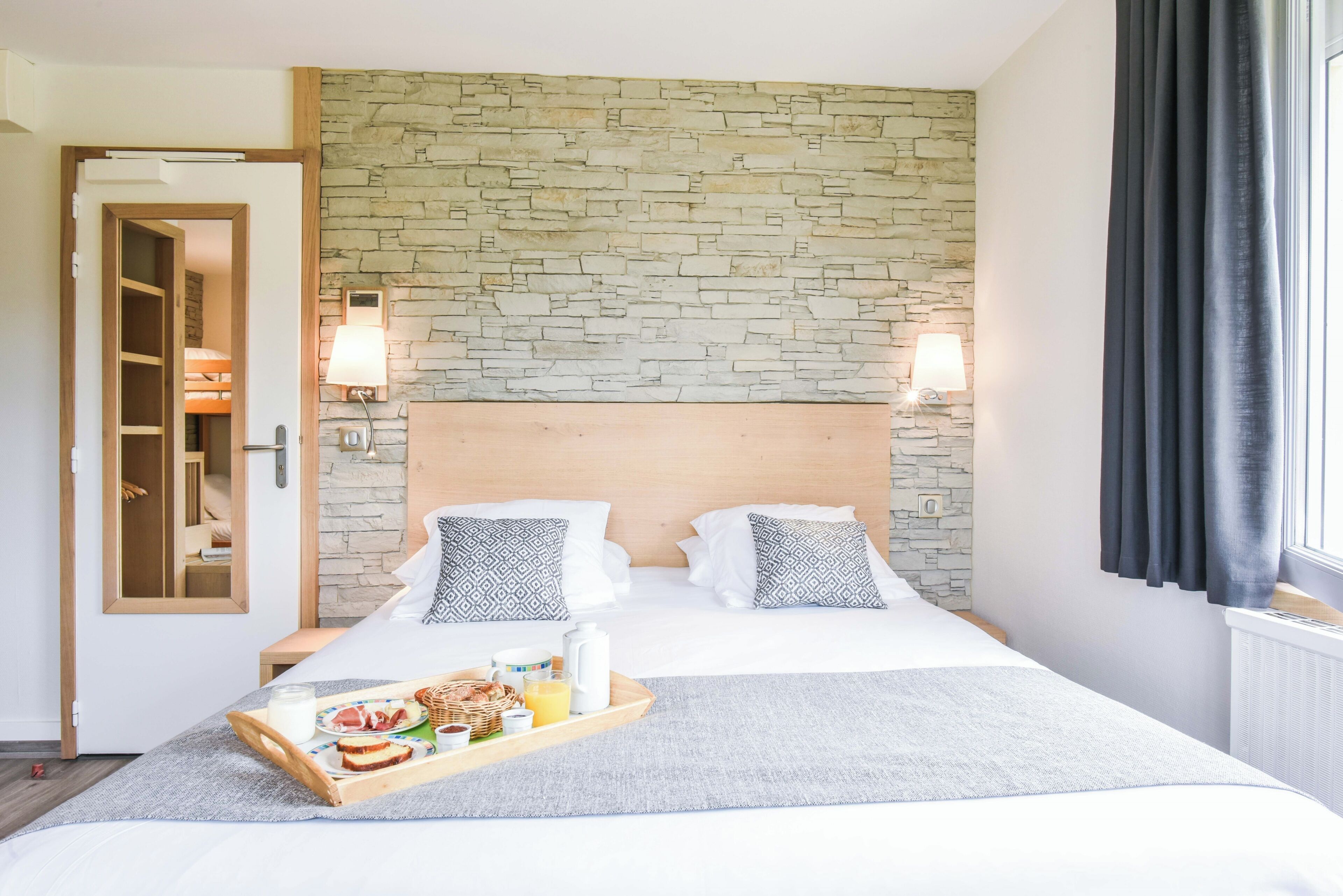 A hotel room with a stone wall, a wooden headboard, two pillows, a tray with breakfast items, and a window with curtains.