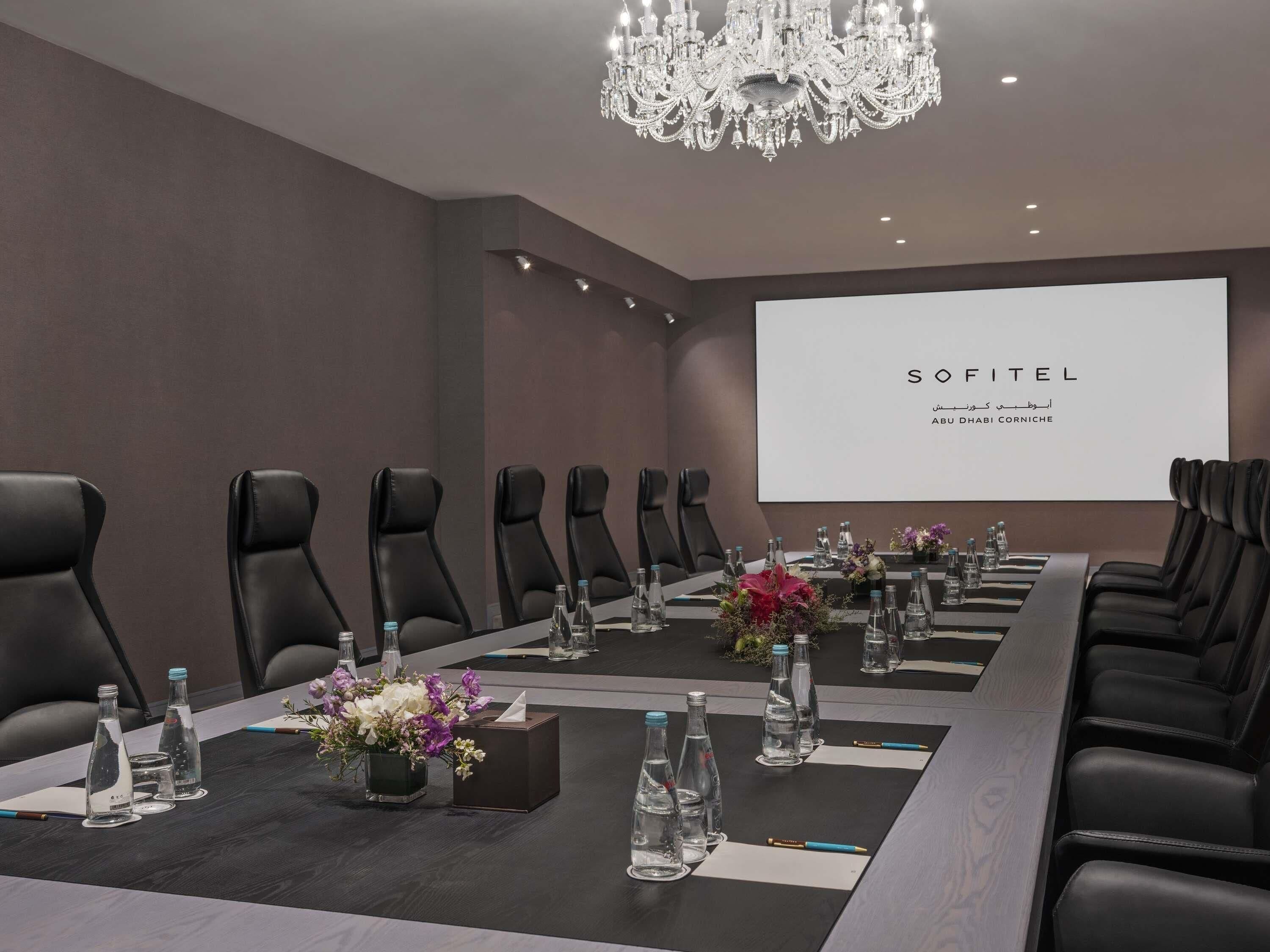 Ein Konferenzraum mit einem langen Tisch, schwarzen Ledersesseln, Wasserflaschen und einer Projektionsleinwand, die 'SOFITEL' zeigt.