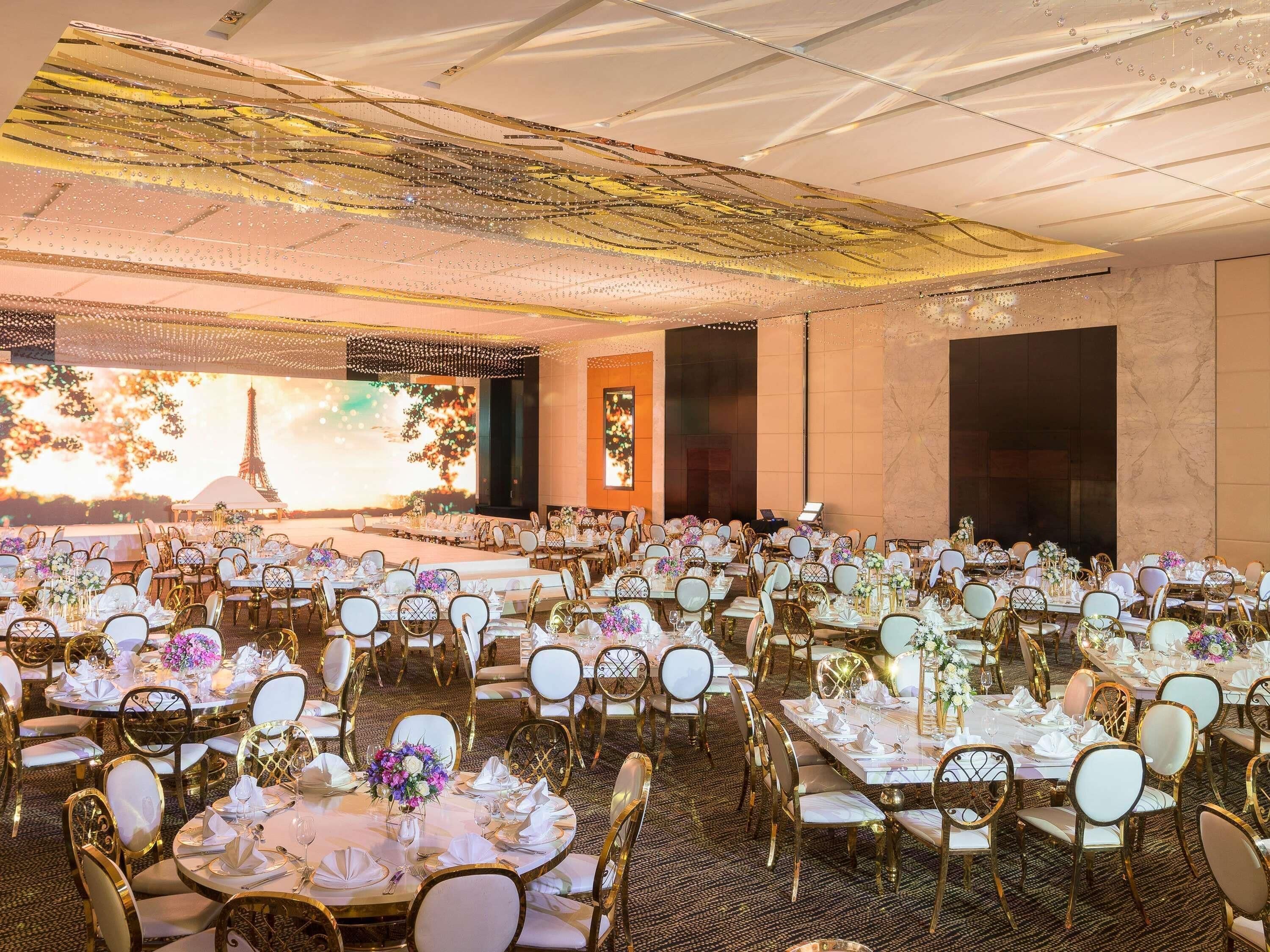 A banquet hall with multiple round tables, each set with white tablecloths, gold chairs, and floral centerpieces.