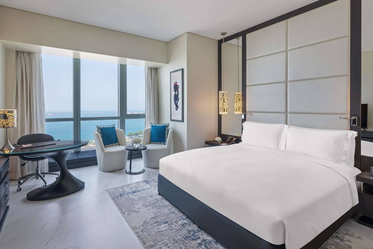 A modern hotel room with a large bed, two armchairs, a desk, and a view of the sea.
