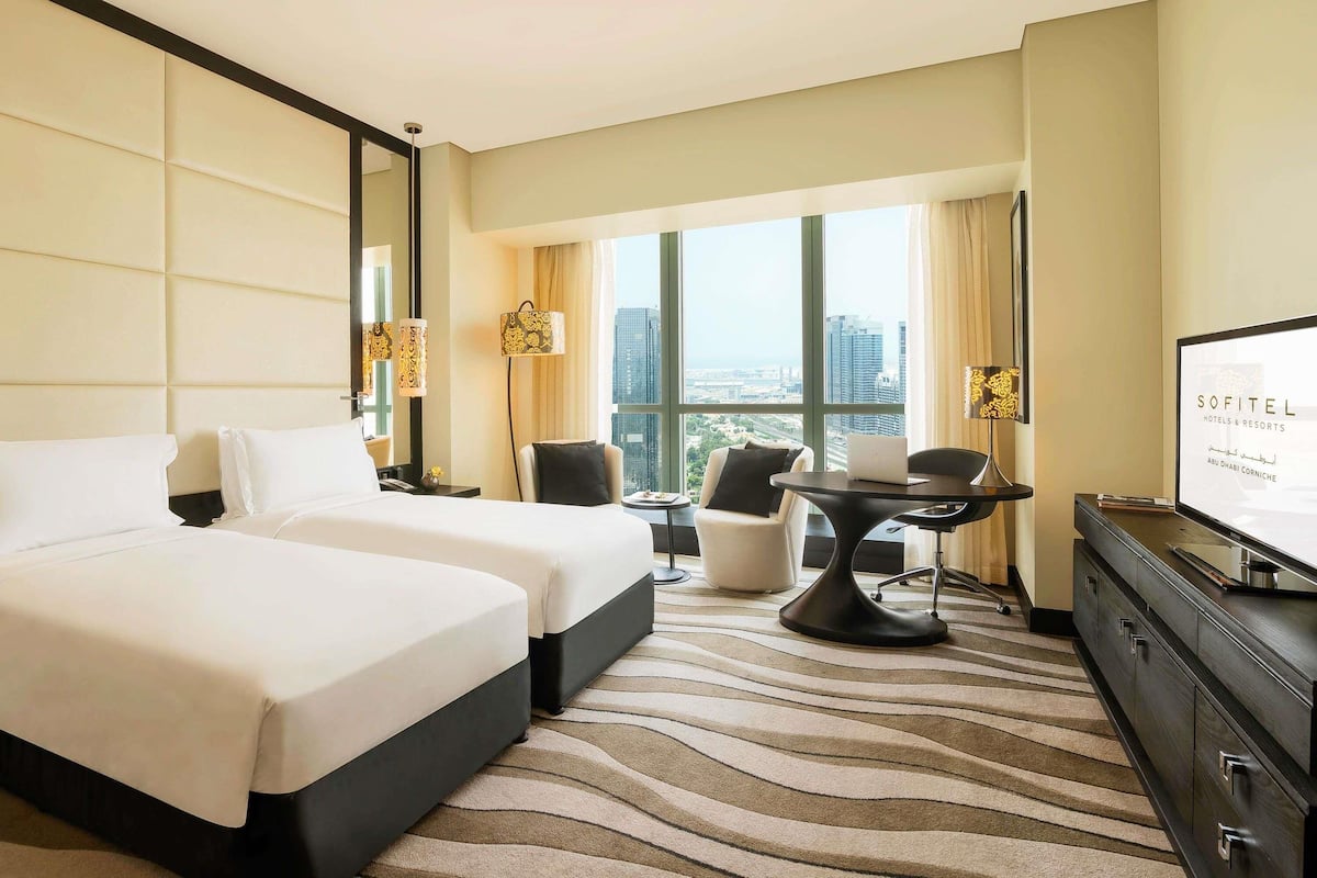A hotel room with two beds, a desk, a chair, a TV, and a large window offering a city view.