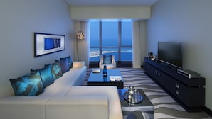 Una habitación de hotel moderna con un ventanal que ofrece vistas al mar, un sofá con cojines estampados, una mesa de centro, un televisor y una nevera pequeña.