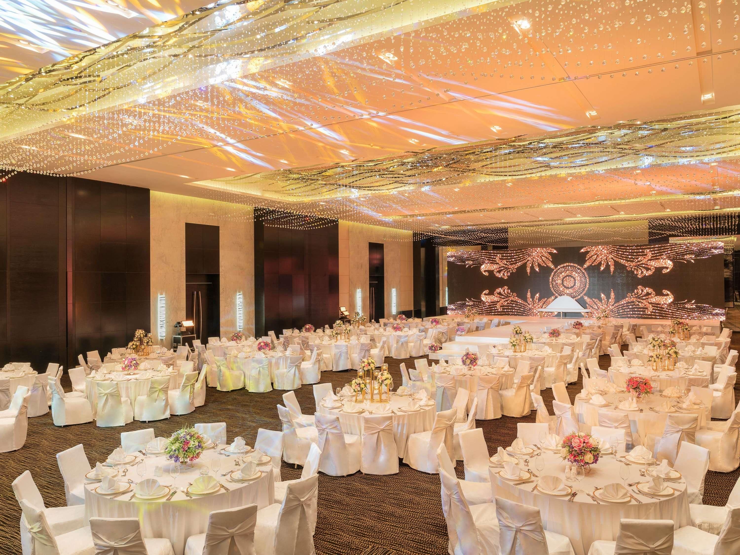 Une salle de banquet avec des tables rondes, des nappes blanches et des chaises, équipée d’un grand écran et d’un éclairage décoratif.