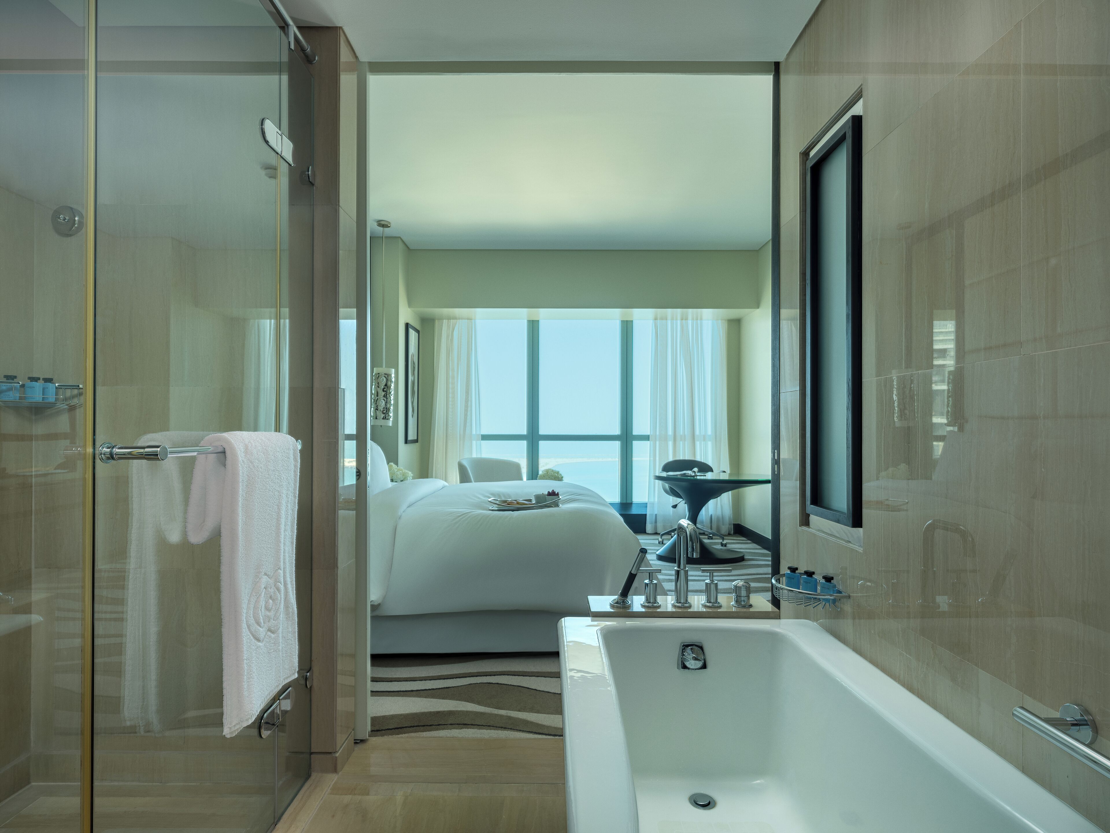 A modern hotel room with a glass shower enclosure, a large bathtub, a bed with white linens, and a view of the city through large windows.
