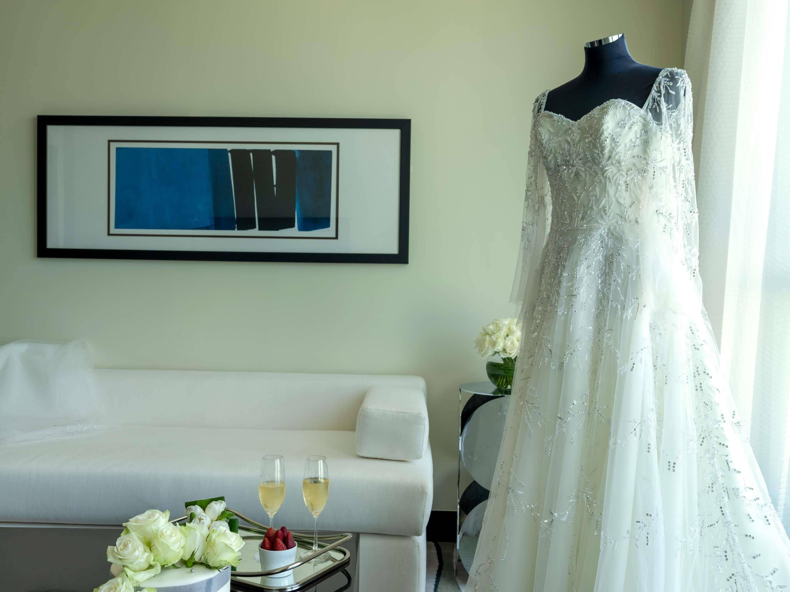 A hotel room with a white sofa, a framed abstract painting, a dress on a mannequin, and a tray with flowers and glasses.