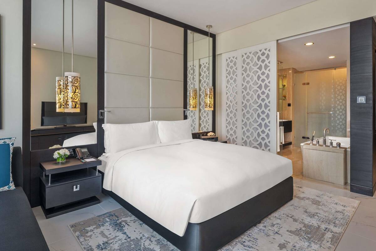 A modern hotel bedroom with a large bed, bedside tables, a TV, and a bathroom visible through an open door.