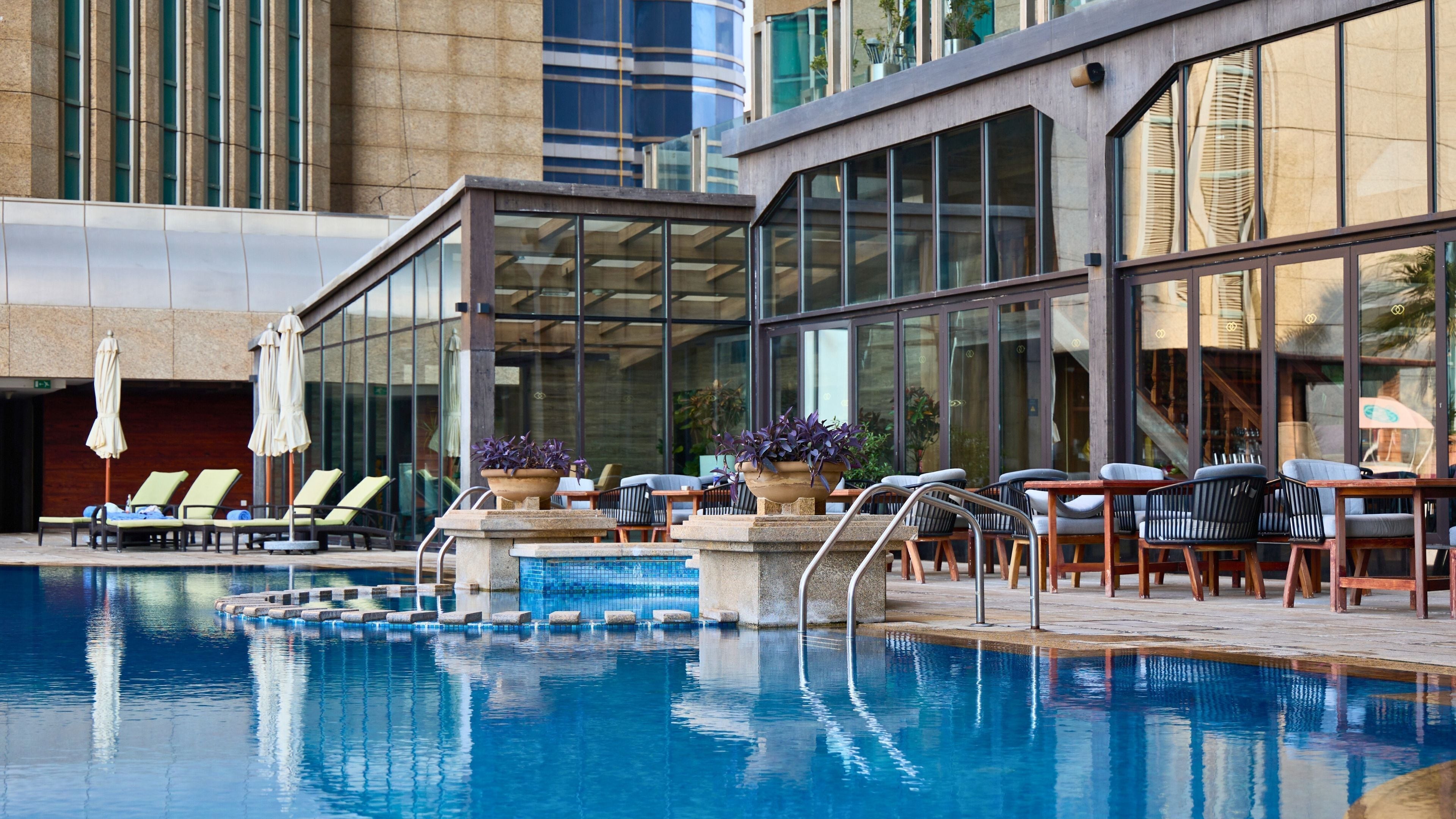 Un área de piscina con sillones reclinables y mesas, rodeada de grandes ventanales y arquitectura moderna.