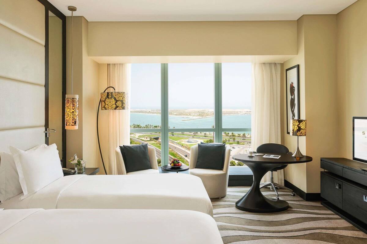 A hotel room with a bed, two chairs, a small table, and a TV. The room has large windows with a view of the sea and a cityscape.