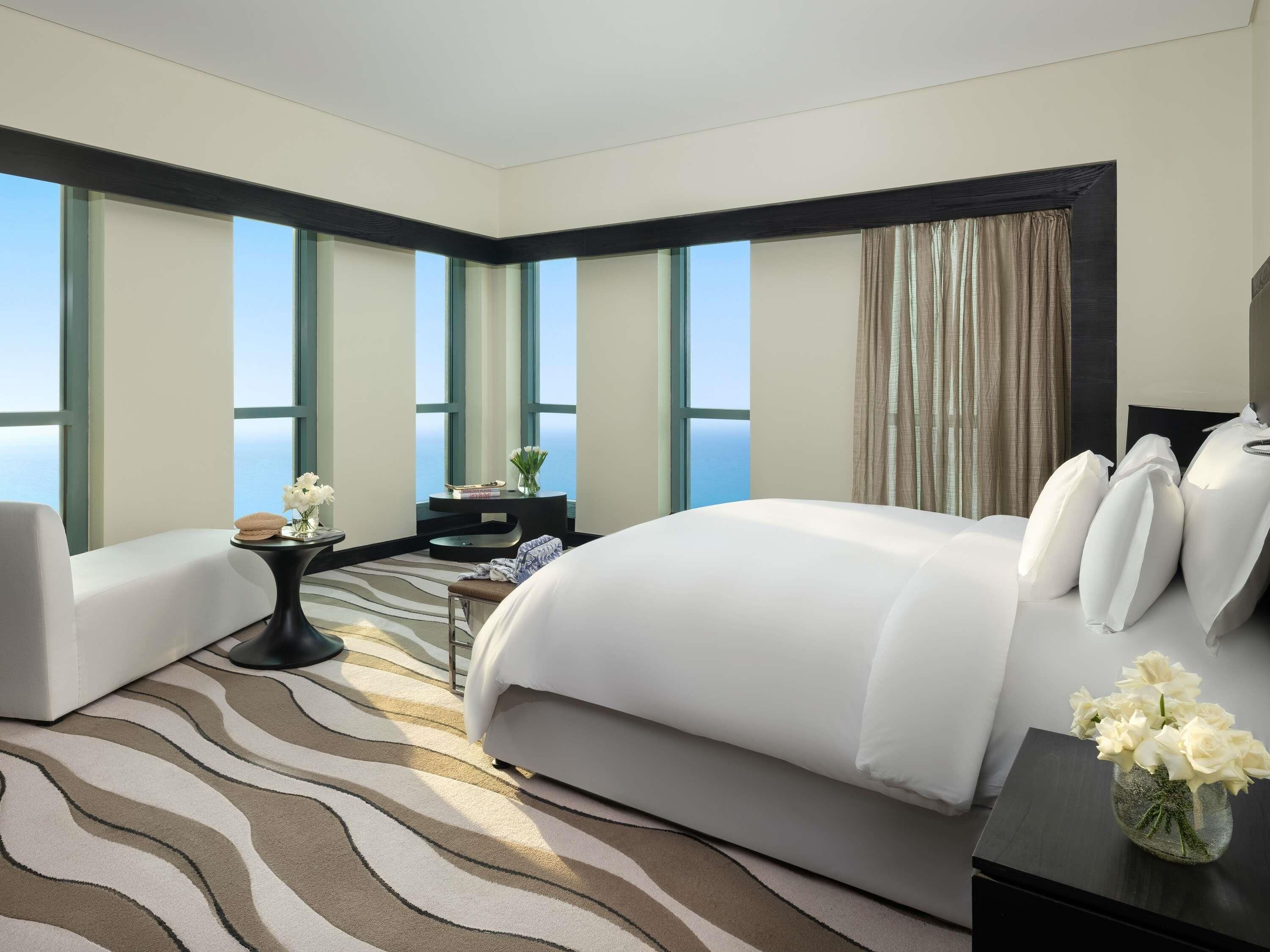 Una habitación de hotel moderna con una cama grande, un área de estar, una mesita con un jarrón de flores y ventanas amplias con vistas al mar.