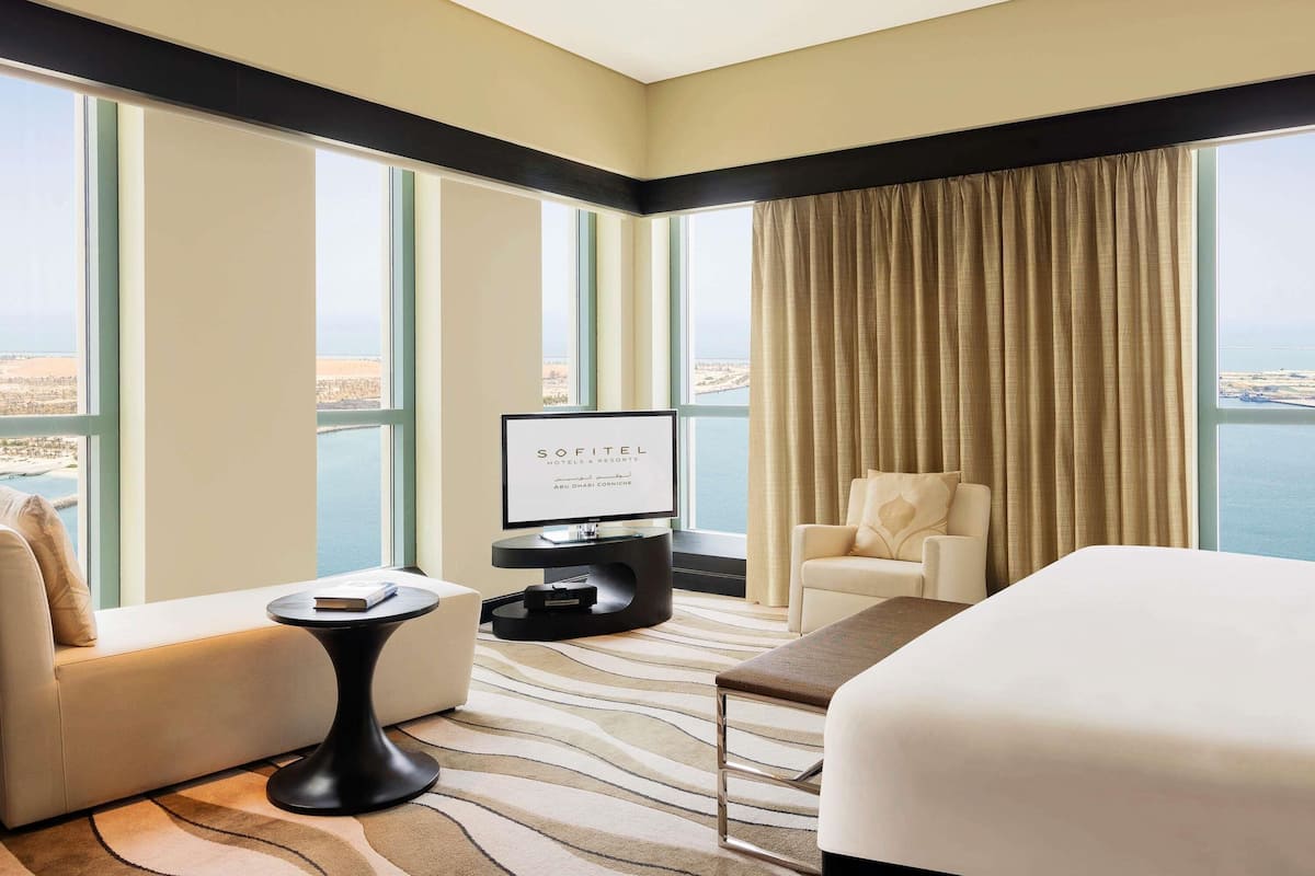 A hotel room with a large bed, a sofa, a small table, a TV, and a view of the sea.
