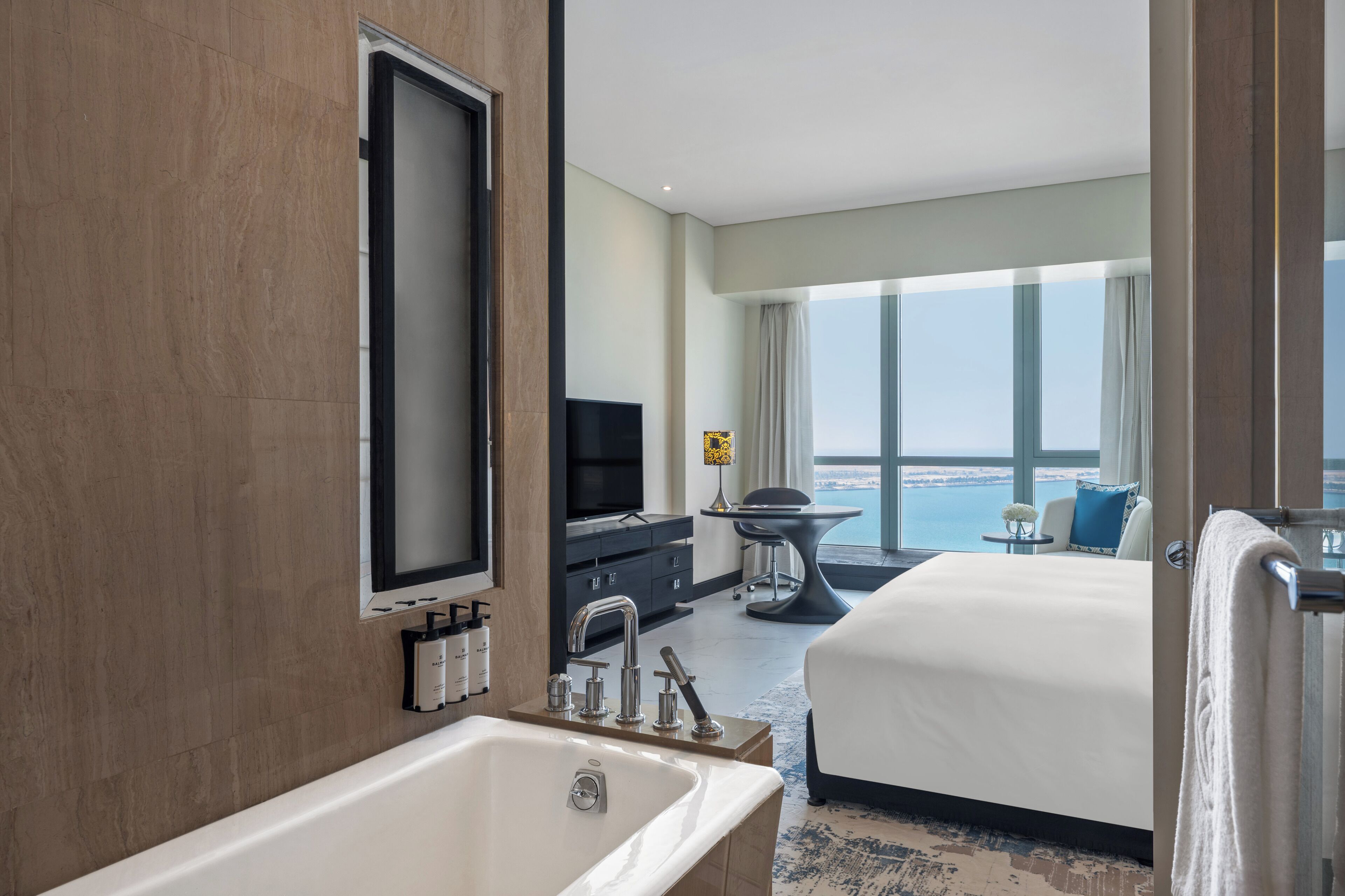 A modern hotel room with a large bed, a sitting area with a view of the sea, a large window, and a bathroom with a bathtub and vanity.