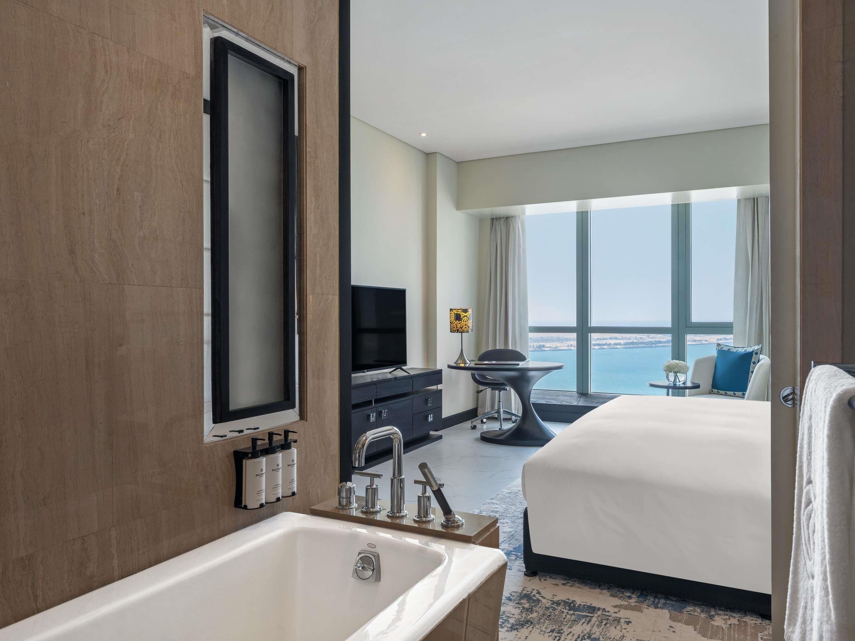 Habitación de hotel con una cama grande, televisión, escritorio con silla, vista al mar y baño con bañera y lavamanos.