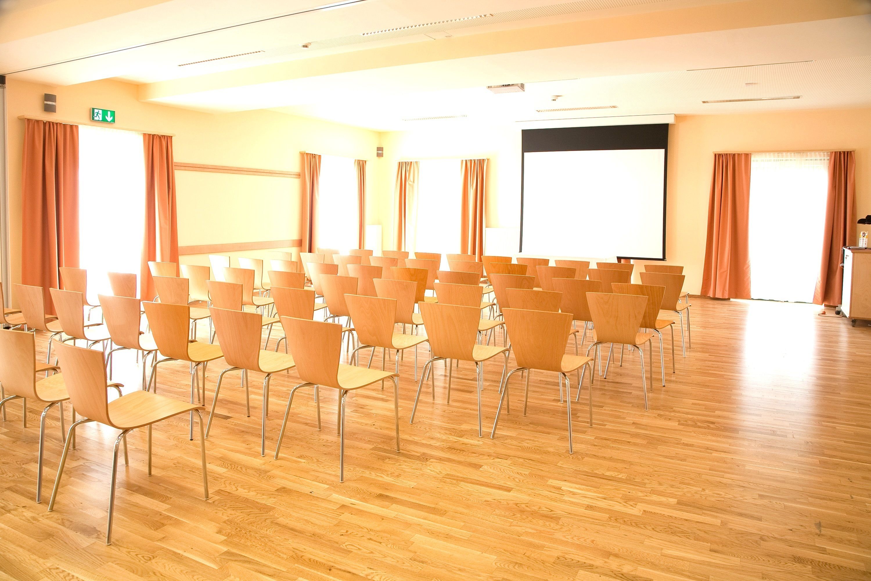 Un salón de conferencias con filas de sillas, una pantalla de proyector y grandes ventanales con cortinas.