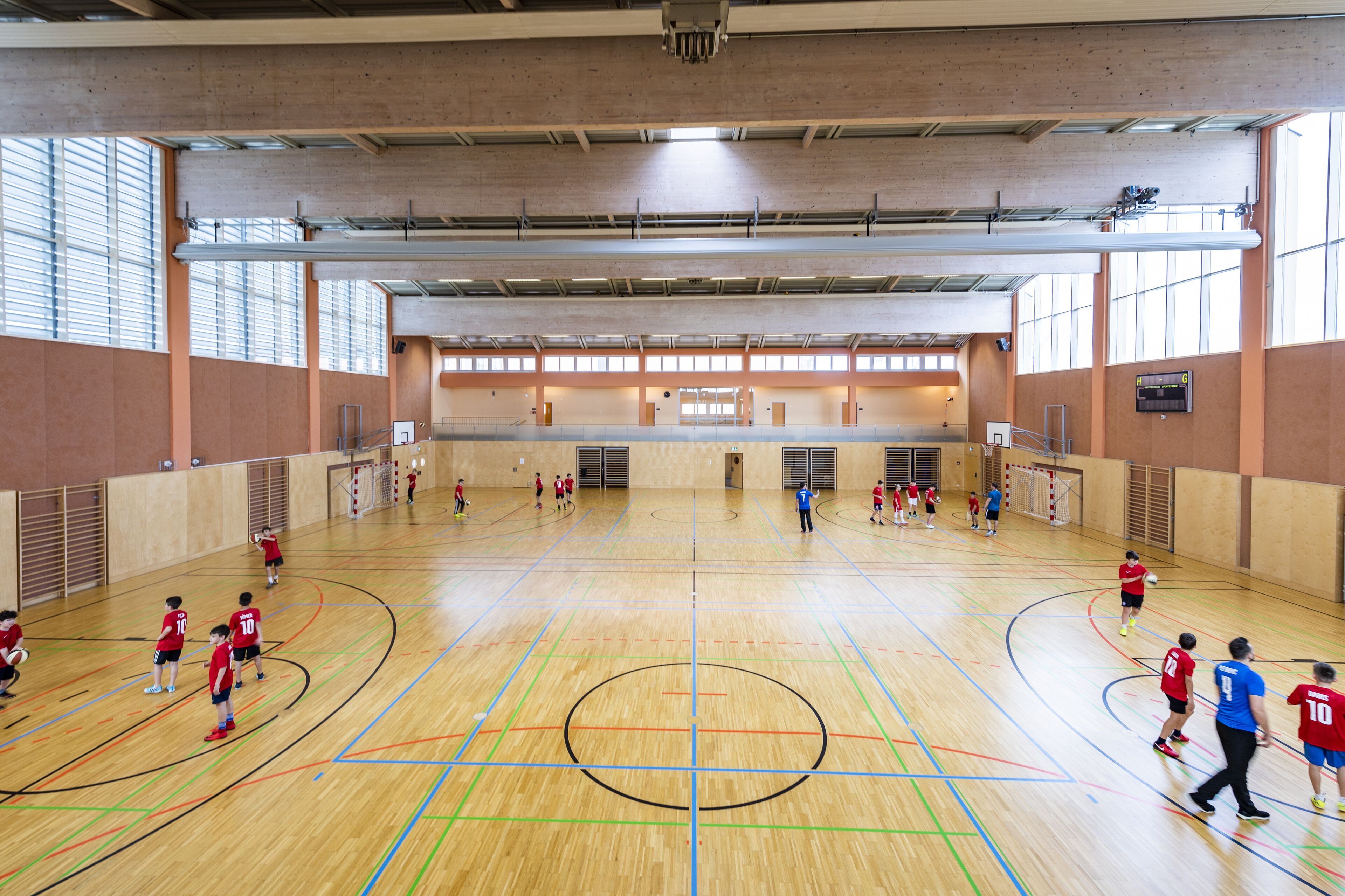 An indoor basketball court with multiple players, a scoreboard, and a large windowed wall.