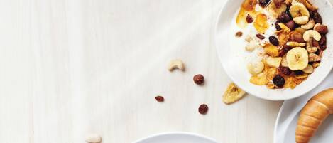 Un desayuno con una fuente de tortitas cubiertas de sirope, un croissant y un bol de cereales con nueces y pasas. También hay dos tazas de té y una servilleta.
