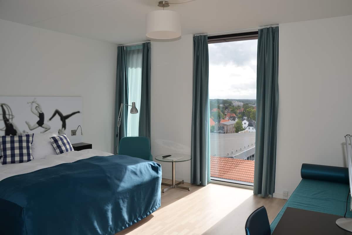 Quarto de hotel moderno com uma cama grande, uma escrivaninha e vista para a cidade através de janelas do chão ao teto.