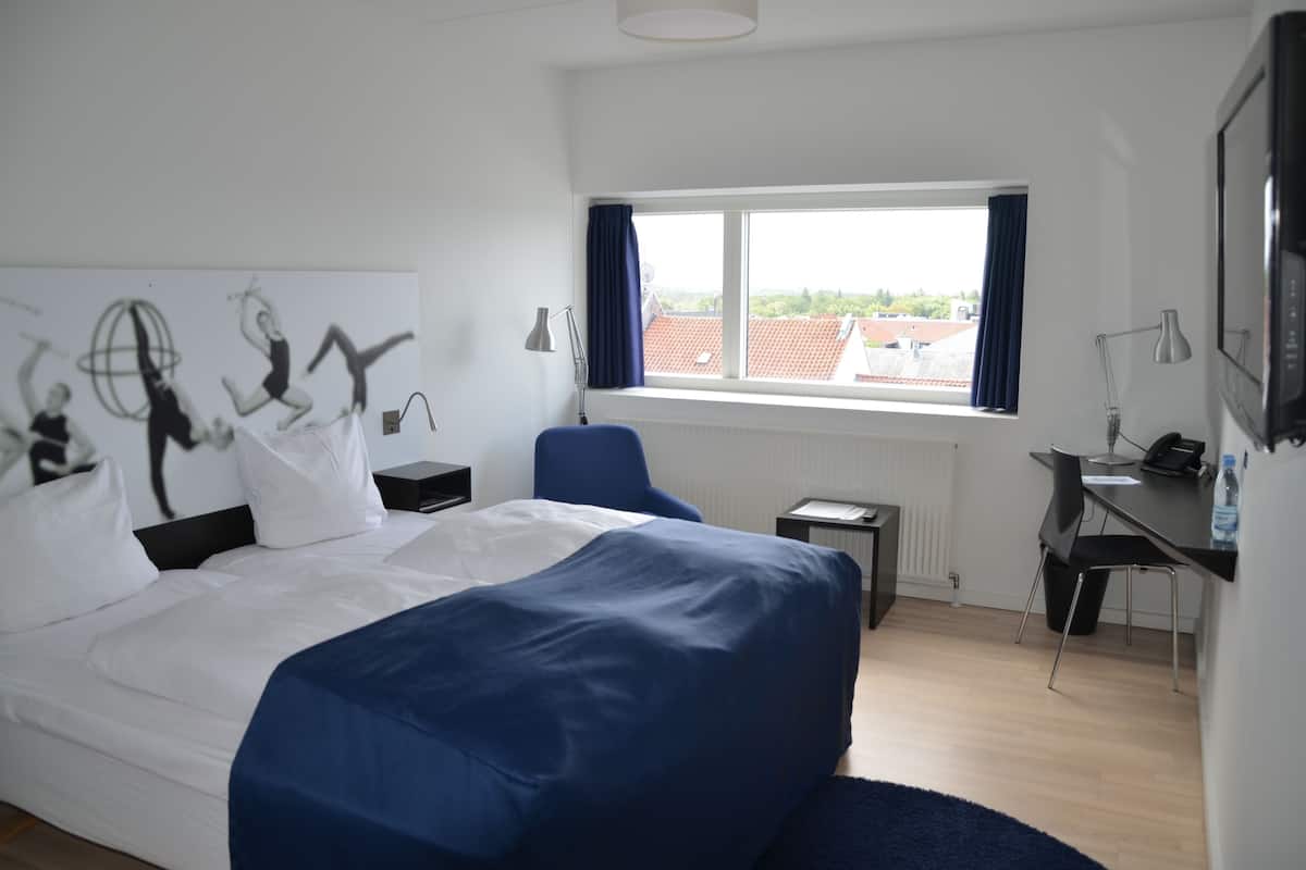 Quarto de hotel com uma cama grande, uma escrivaninha, uma cadeira, uma janela com vista para edifícios e uma parede decorada com obras de arte.