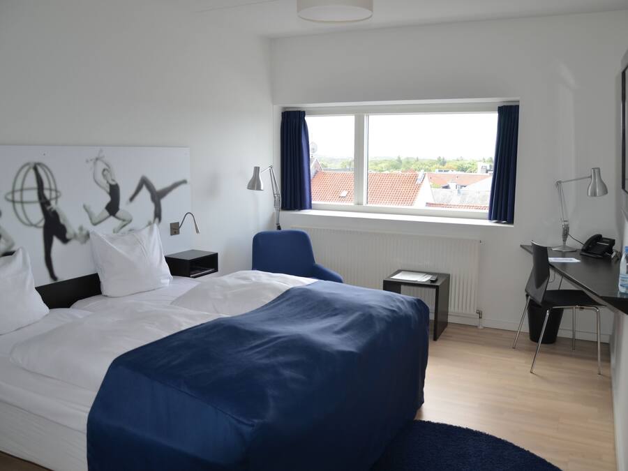 Une chambre d’hôtel avec un grand lit, un bureau, une chaise, une fenêtre donnant sur des immeubles et un mur orné d’œuvres d’art.