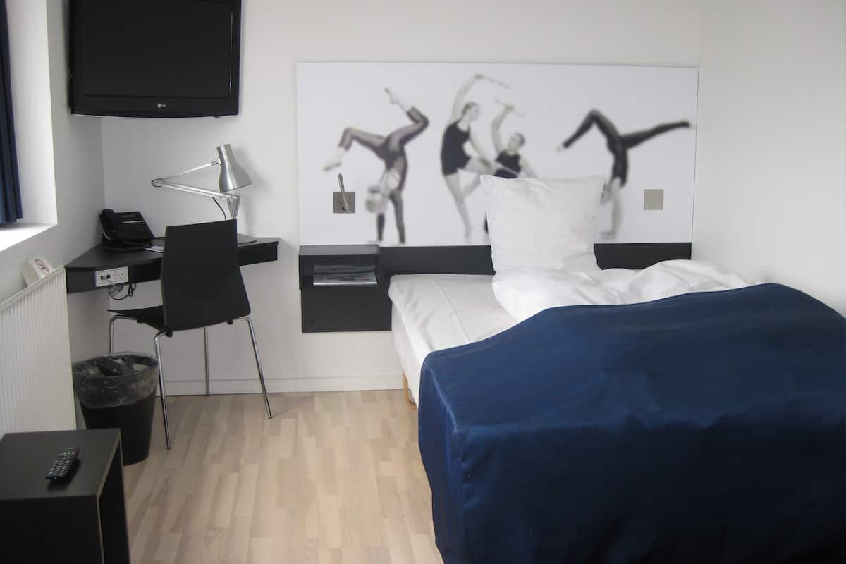 Quarto de hotel com uma cama, uma escrivaninha com cadeira, uma TV na parede e uma obra de arte retratando dançarinos.