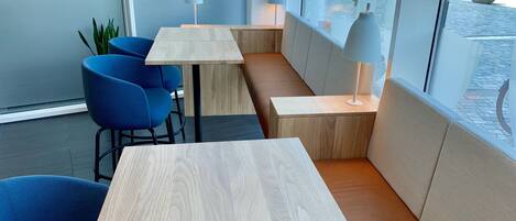 A modern interior with wooden tables and blue chairs, large windows offering a view of the street.