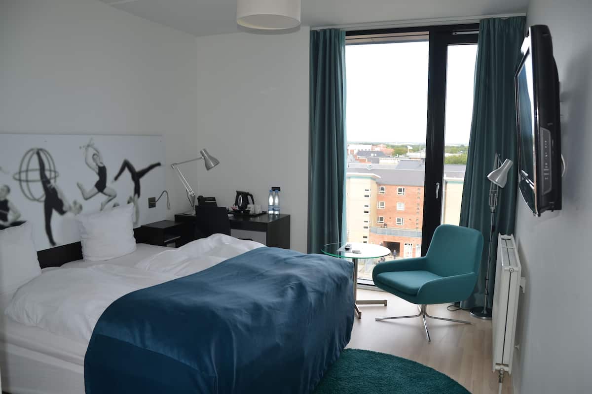 Quarto de hotel moderno com uma cama grande, uma escrivaninha, uma cadeira e vista para a cidade pela janela.