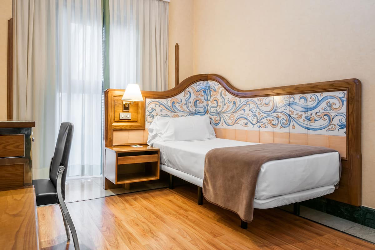 A hotel room with a wooden bed, a nightstand with a lamp, a desk, and a chair.