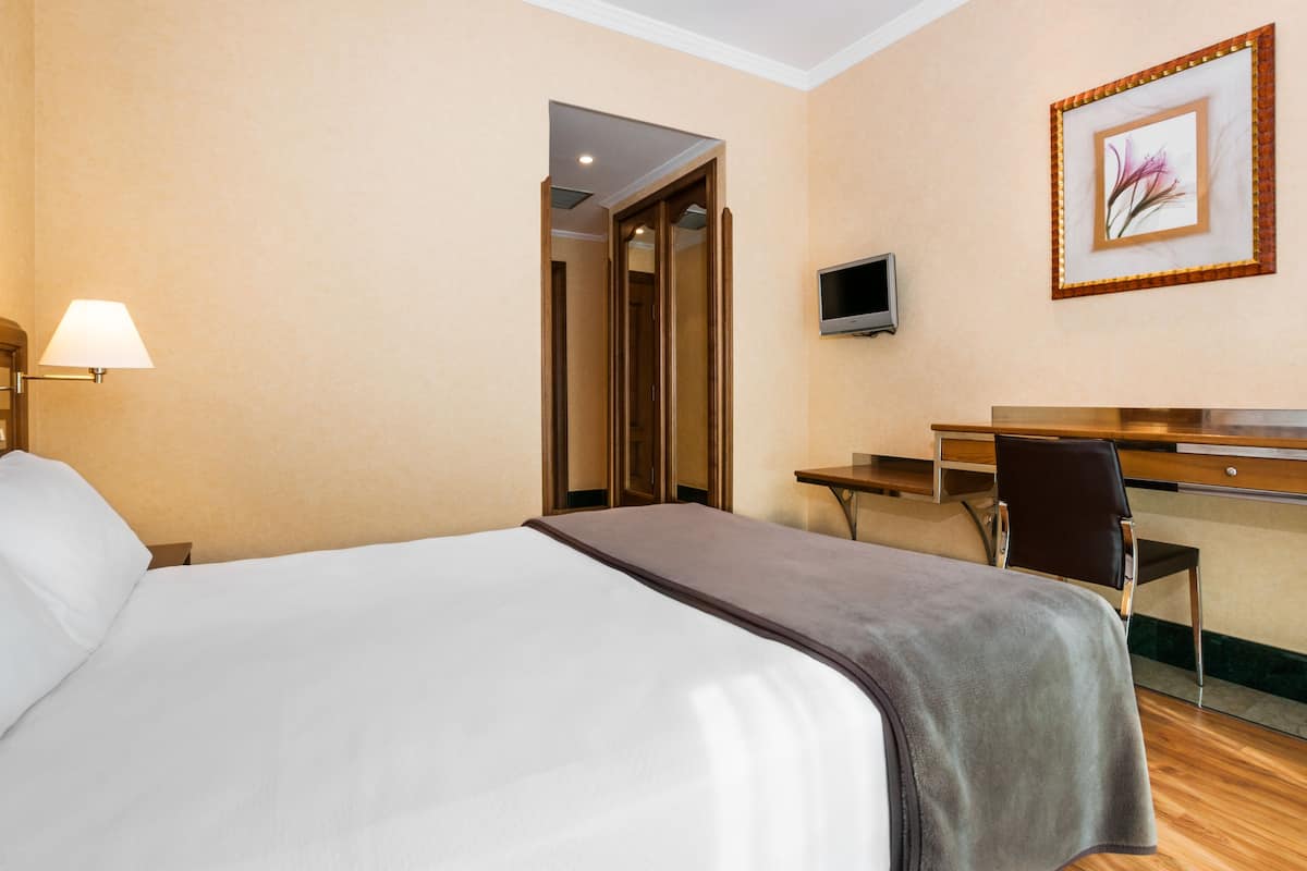 A hotel room with a bed, a desk, a chair, a TV, and a door leading to another room.