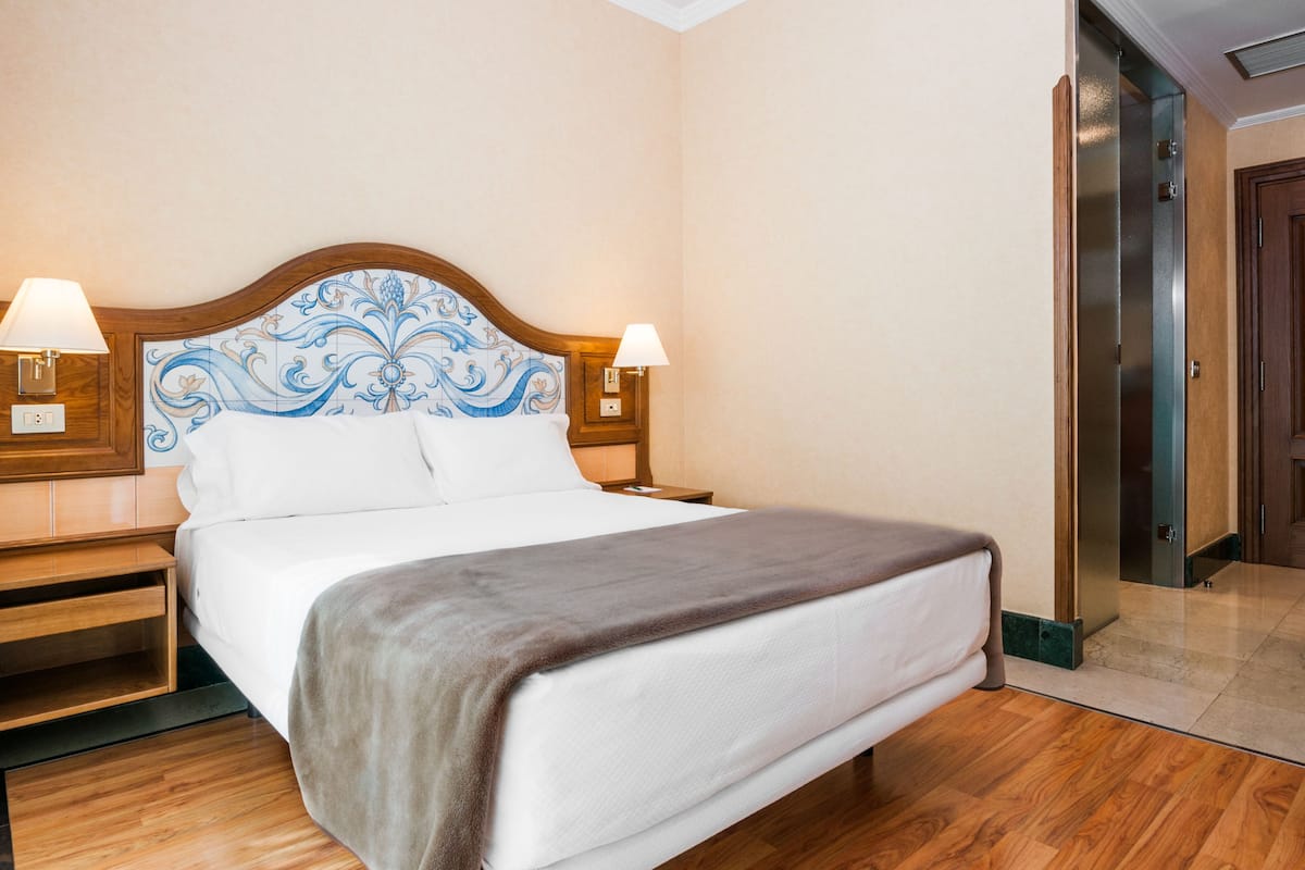 A hotel room with a large bed, wooden headboard, two bedside lamps, and a door leading to another room.