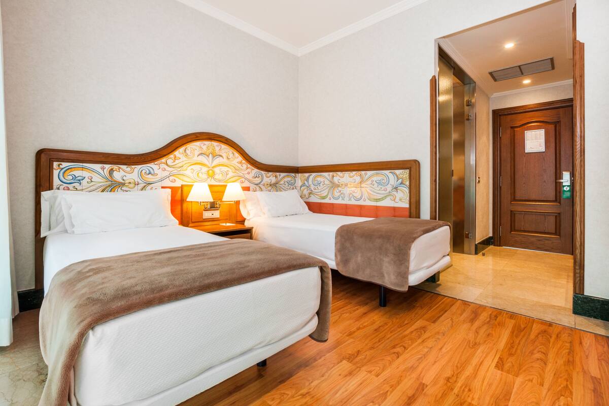 A hotel room with two beds, a wooden headboard, a nightstand with a lamp, and a wooden door.
