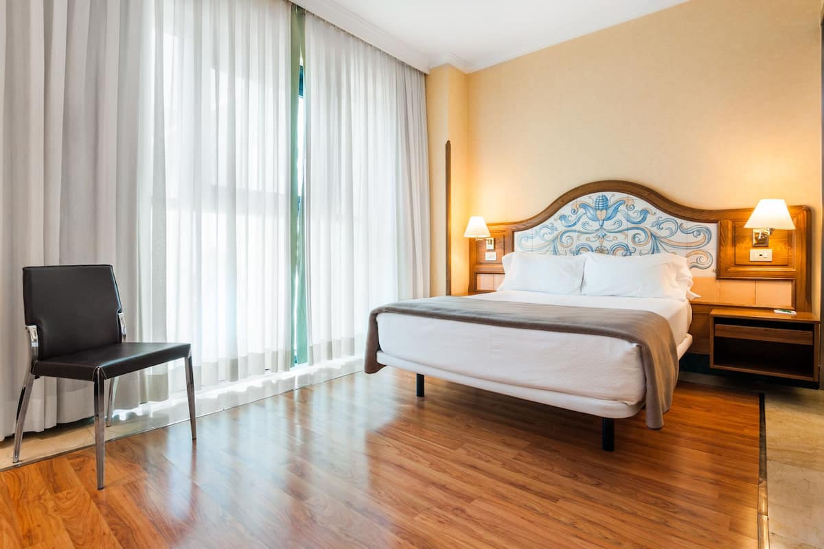 A hotel room with a bed, two bedside tables, a chair, and a large window with sheer curtains.