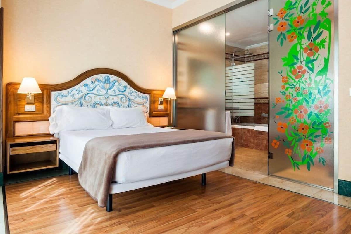 A hotel room with a bed, two bedside lamps, a wooden headboard, a sliding glass door with a floral mural, and a wooden floor.