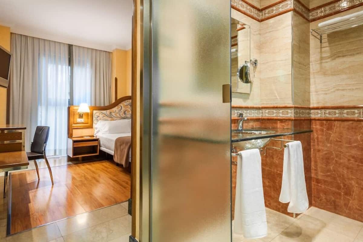 A hotel room with a bed, bedside table, lamp, chair, and a bathroom with a glass shower enclosure and towel rack.
