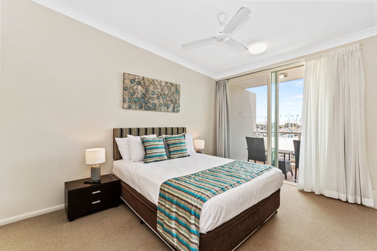 A hotel room with a large bed, a ceiling fan, a nightstand with a lamp, and a view of a balcony with a cityscape.