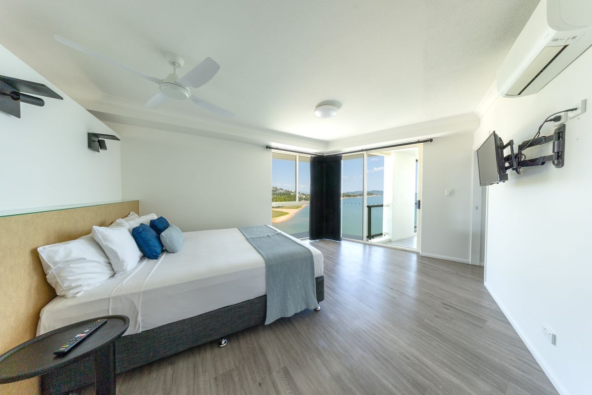 A modern hotel room with a bed, a remote control, a ceiling fan, and a large window offering a view of the outdoors.