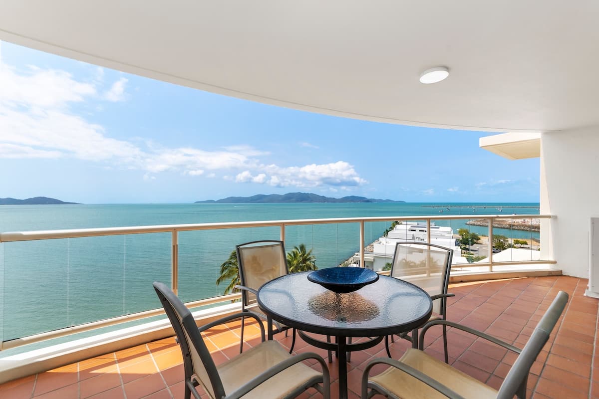 A balcony with a table and chairs, overlooking a body of water and distant mountains.