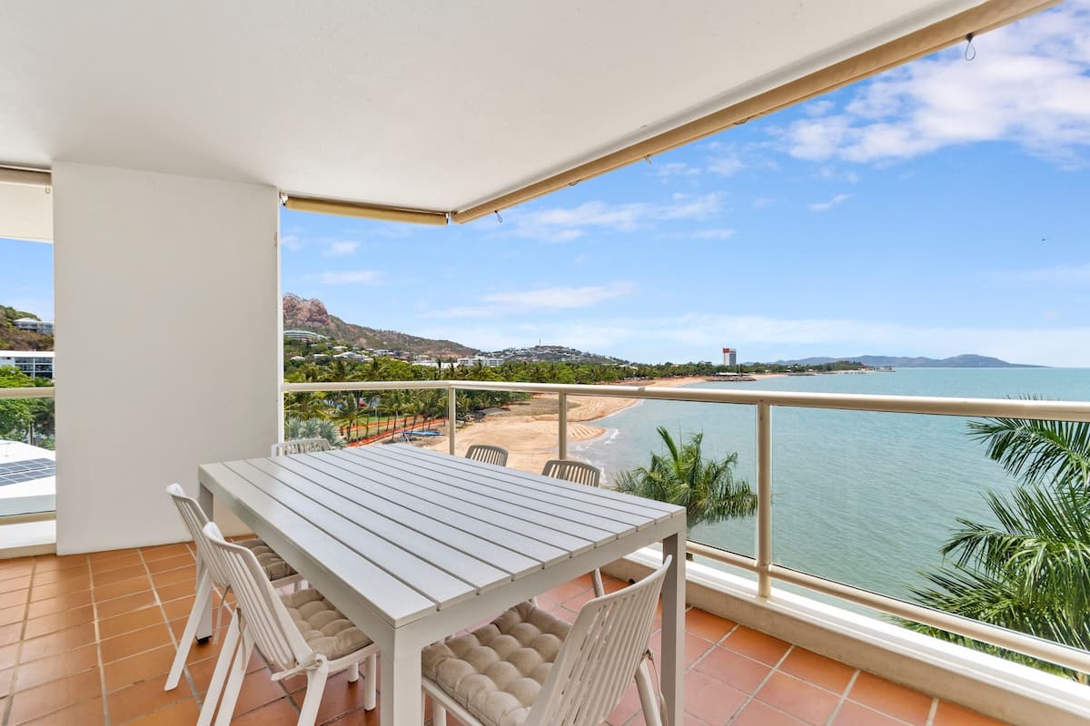 A balcony with a table and chairs, overlooking a beach and ocean with distant hills.