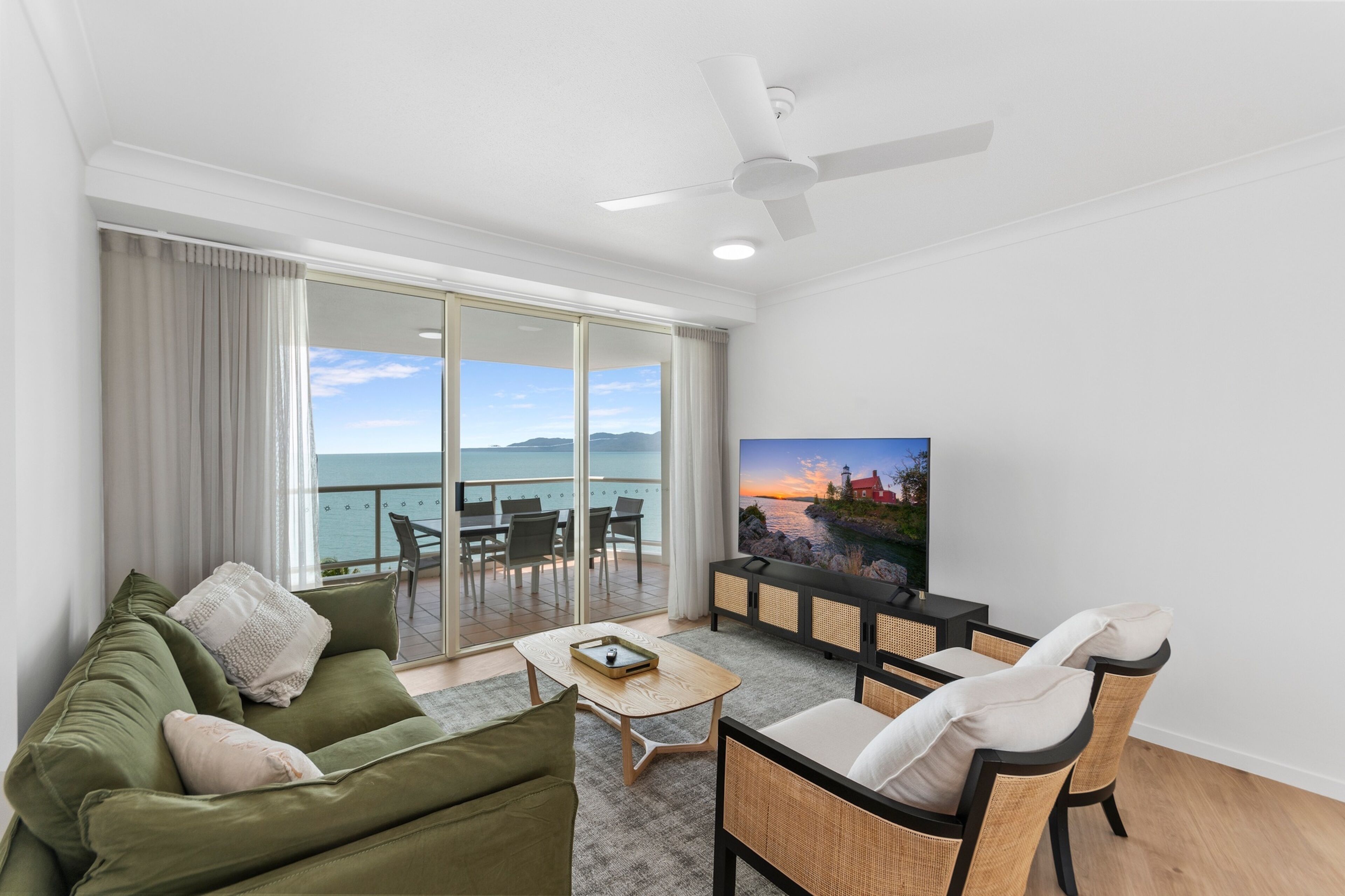 A modern living room with a green sofa, a coffee table, and a TV on a stand. There is a balcony with a view of the sea.