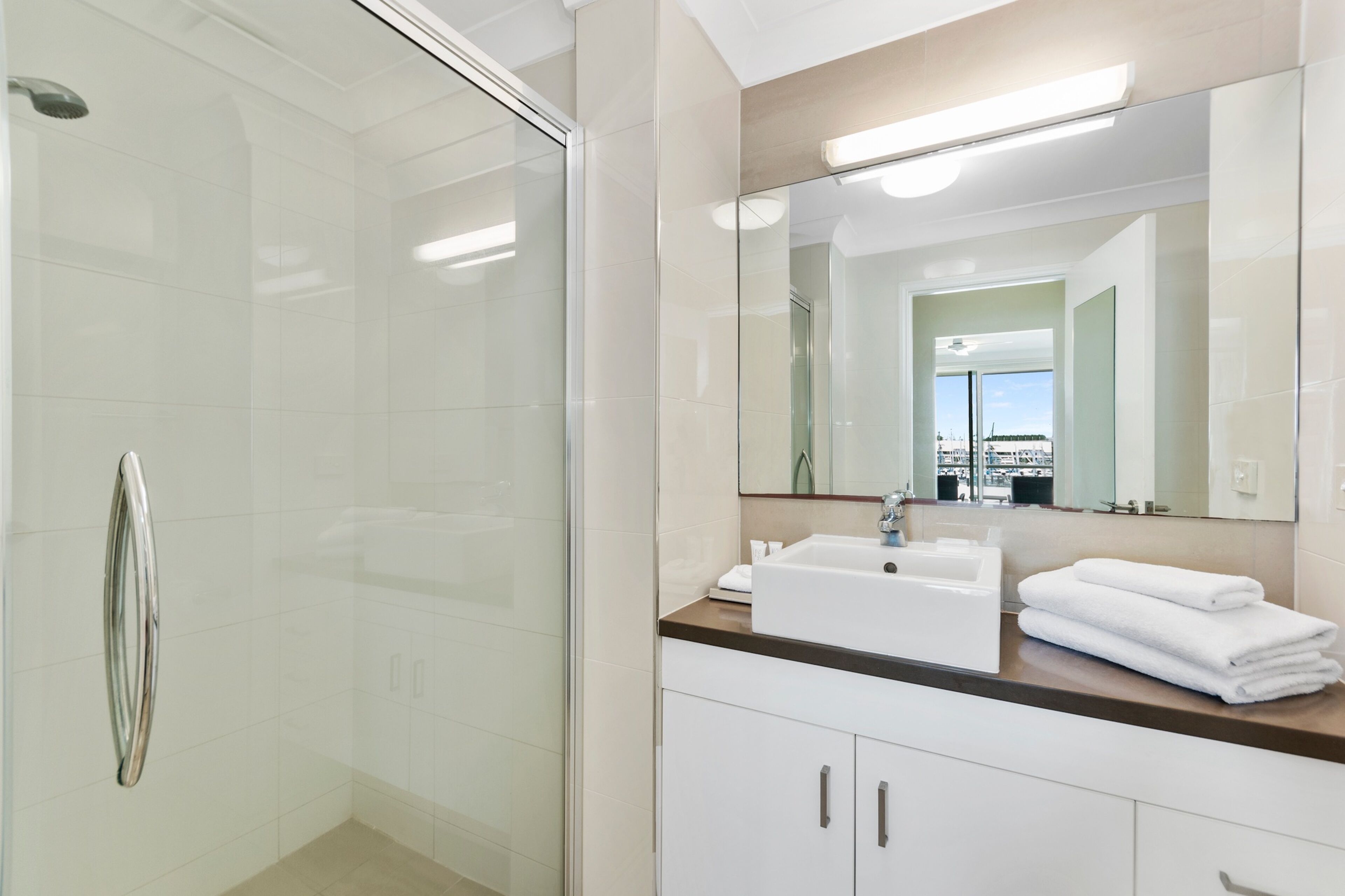 A modern bathroom with a glass shower enclosure, a white sink with a mirror above it, and a view of the city through the window.