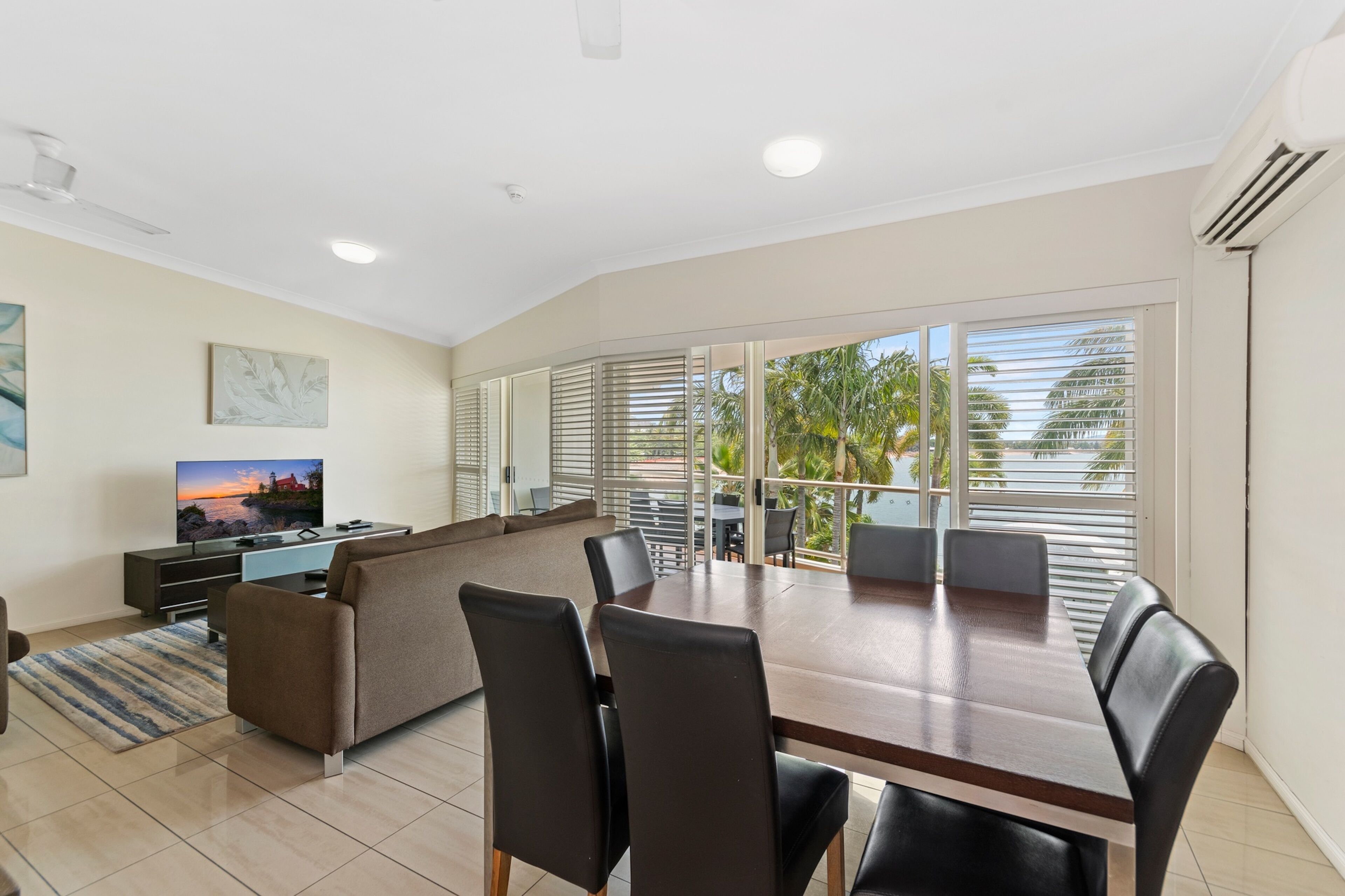 A modern living room with a dining area, a sofa, a TV, and a balcony with palm trees.