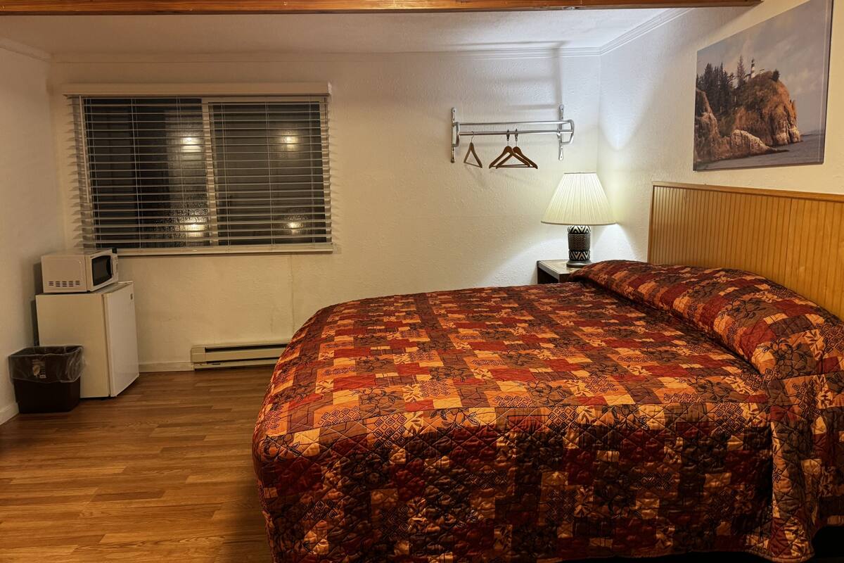 Une chambre avec un plafond en bois, un lit recouvert d’une courtepointe à motifs, une petite table de chevet avec une lampe, une fenêtre avec des stores et un réfrigérateur.